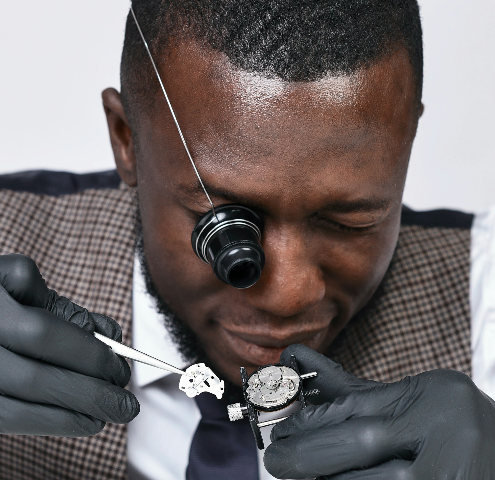 A watchmaker in a houndstooth jacket examining a movement through a loupe